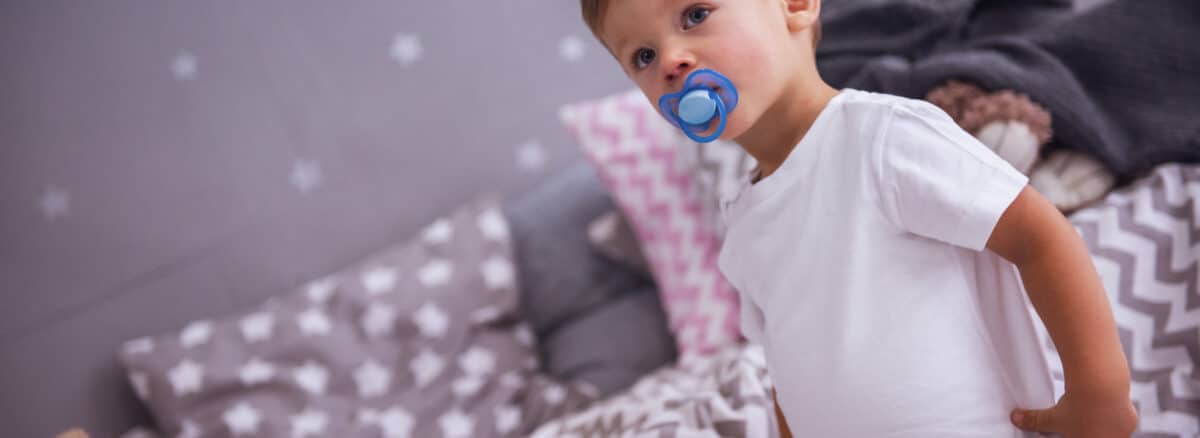 A cute little boy with a pacifier that's standing in in his bedroom