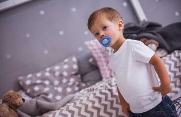 A cute little boy with a pacifier that's standing in in his bedroom