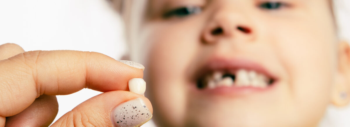 Fingers holding a baby tooth with little girl face looking at tooth and showing teeth in the background.