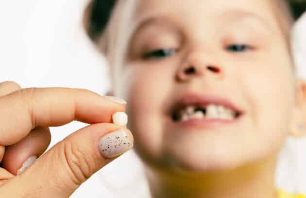 Fingers holding a baby tooth with little girl face looking at tooth and showing teeth in the background.